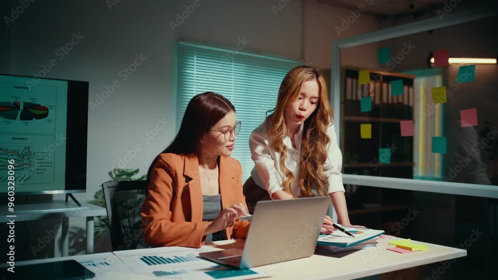 Two young businesswomen are working late in their office, collaborating on a project using a laptop. One of them is showing something on the laptop screen to her colleague Two young businesswomen are working late in their office, collaborating on a project using a laptop. One of them is showing something on the laptop screen to her colleague