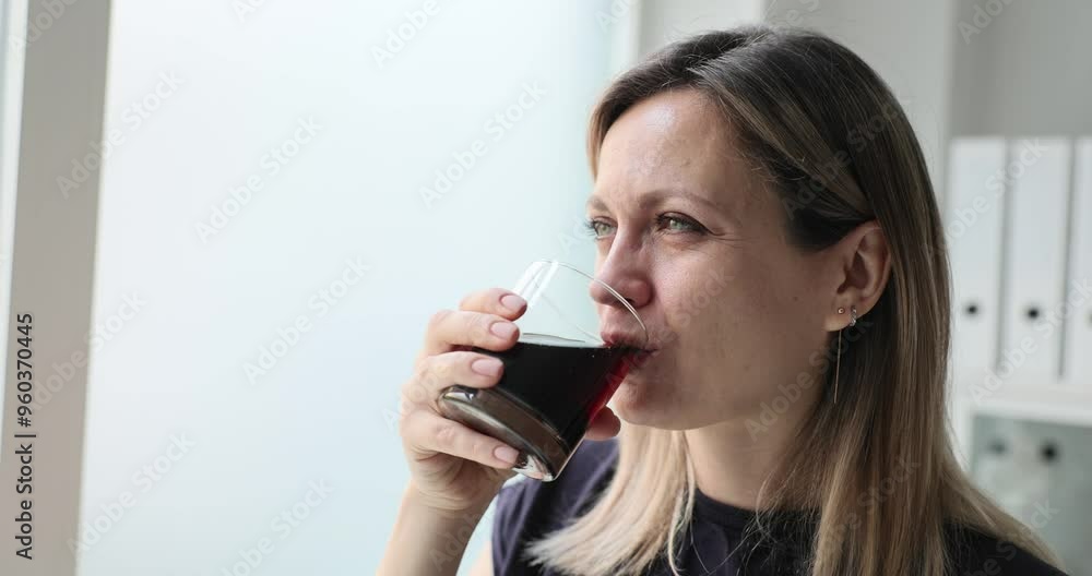Woman holds glass drinking sweet beverage against window. Female with delightful smile enjoys fresh taste of delicious beverage in office Woman holds glass drinking sweet beverage against window. Female with delightful smile enjoys fresh taste of delicious beverage in office