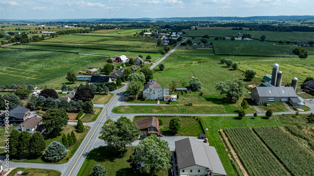 This view showcases a serene rural community with houses and farms nestled among lush green fields under a clear blue sky, embodying the tranquility of summertime. This view showcases a serene rural community with houses and farms nestled among lush green fields under a clear blue sky, embodying the tranquility of summertime.