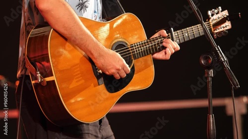 Close-up of a guitarist’s hands energetically strumming the strings of his guitar. The stage lights create a vibrant atmosphere, enhancing the dynamic performance.