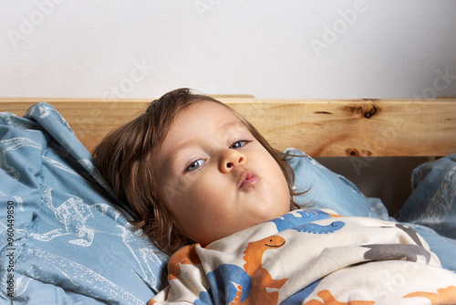Niño rubio de dos años echado en su cama azul, él está mirando a la cámara, está haciendo una mueca. Cubrecama azul. Interior