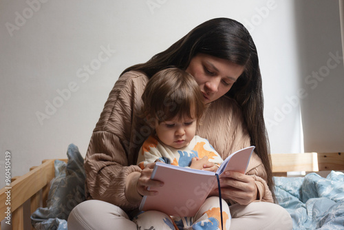 Bild auf Leinwand Mamá con bebé de un año sentado en sus piernas, ella le está leyendo un cuento, ellos están sentados sobre una cama