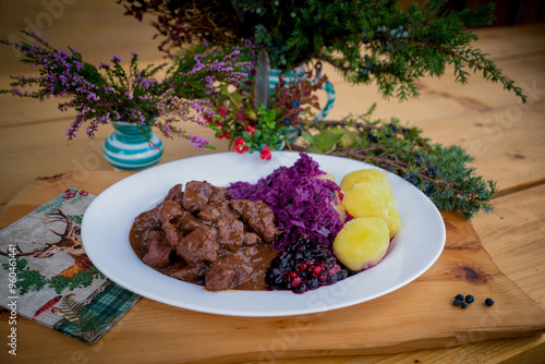 ready to eat and served on a white plate on a wood table - goulash from venison meat with potatoes, red cabbage and craneberries -  cooked on a charcoal grill at outdoor photo