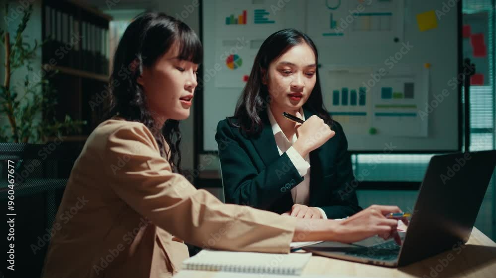 Two businesswomen are working late in the office. They are discussing charts and graphs on a laptop. One of them is wearing a headset Two businesswomen are working late in the office. They are discussing charts and graphs on a laptop. One of them is wearing a headset