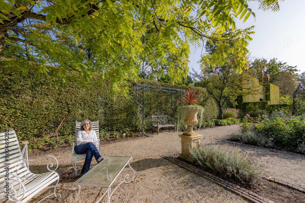 Adult woman sitting comfortably on metal chair in inner gardens of Arcen castle, lush bushes and trees, casual clothing, white blouse and jeans, sunny spring day in Limburg, Netherlands Adult woman sitting comfortably on metal chair in inner gardens of Arcen castle, lush bushes and trees, casual clothing, white blouse and jeans, sunny spring day in Limburg, Netherlands