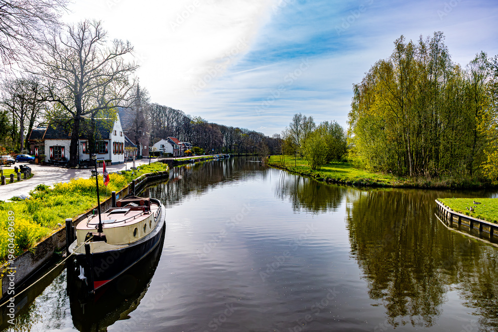 Boat anchored on canal bank between houses and bare trees against blue sky in background, rural vehicular road along river, sunny spring day in Oud-Zuilen, Utrecht, Netherlands Boat anchored on canal bank between houses and bare trees against blue sky in background, rural vehicular road along river, sunny spring day in Oud-Zuilen, Utrecht, Netherlands