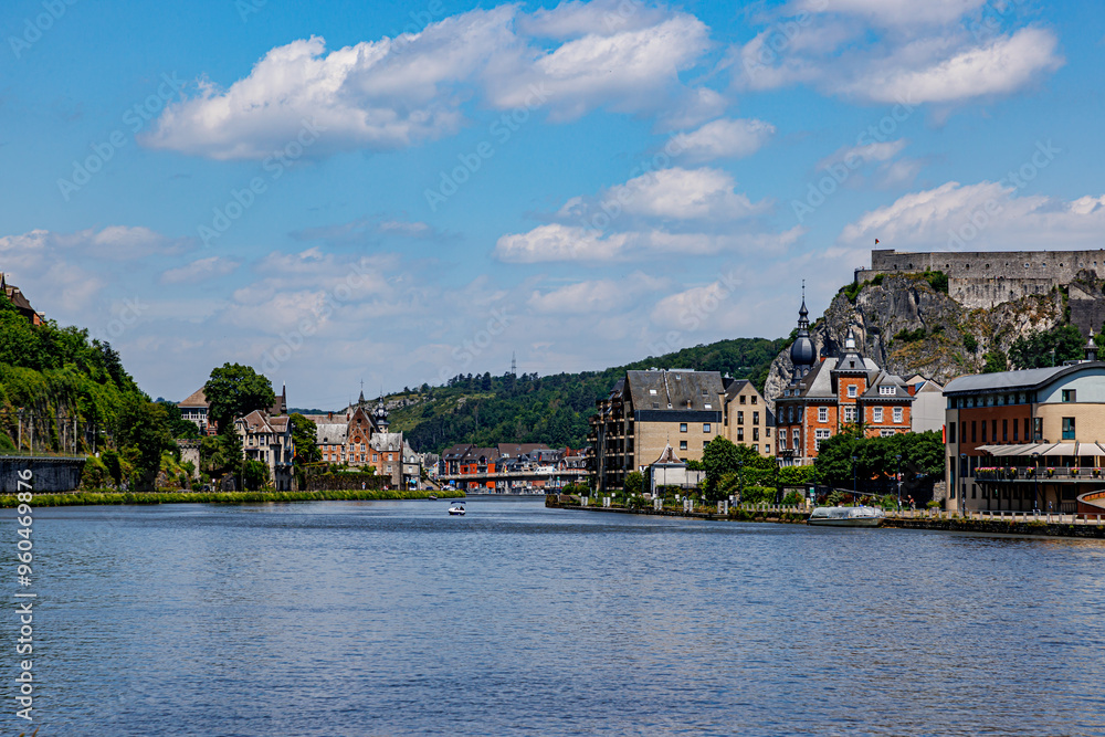 Obraz premium View of Meuse river with cityscape of Dinant resort town and citadel on top of rocky mountain against blue sky in background, lush trees on hill, sunny summer day in Namur province, Wallonia, Belgium