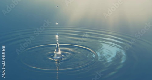 A single drop of water falls into a calm, mirror-like surface of the sea, creating gentle concentric ripples and a soft splash. The serene scene captures the peaceful interaction of water and motion, 