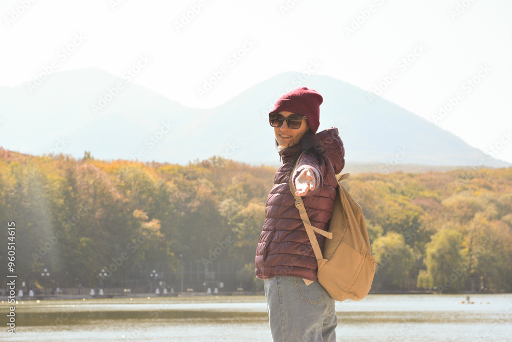 A beautiful woman with a backpack behind her back smiles at the camera and rests on the pier of a lake in the mountains. Vacation concept