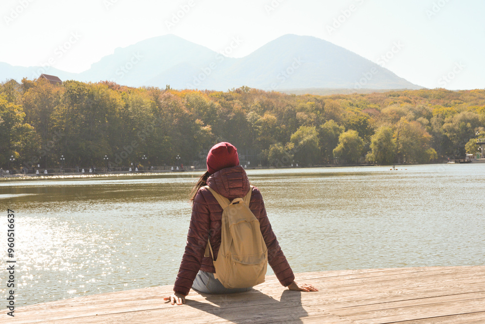 A beautiful tourist woman with a backpack behind her is resting on the lake pier. Vacation concept