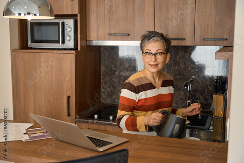 Senior woman having break and making tea while working with laptop in hotel