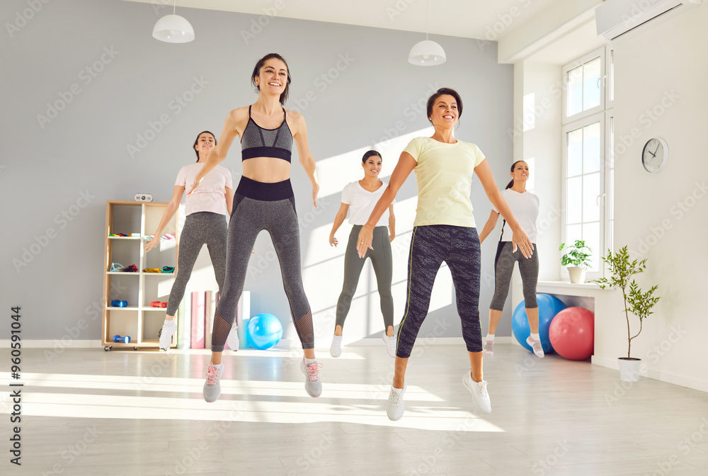 Fototapeta premium Group of happy cheerful beautiful sporty young women in sports clothes doing jumping exercises all together during an active fitness workout at the gym