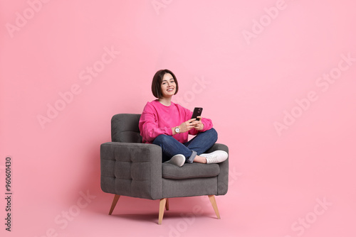 Smiling woman with smartphone sitting on armchair against pink background