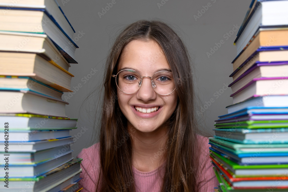 Smiling girl surrounded by stacks of books wearing glasses. A cheerful young girl with long hair and glasses smiles brightly while sitting between two large stacks of books. Smiling girl surrounded by stacks of books wearing glasses. A cheerful young girl with long hair and glasses smiles brightly while sitting between two large stacks of books.