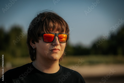 A young man wearing reflective sunglasses and a cross earring gazes into the distance while standing outdoors. His sunglasses reflect the golden hues of the sunset.