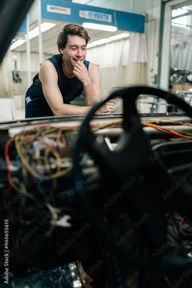 Fototapeta premium Vertical shot of cheerful mechanic smiling while thinking about how to repair electrical system of a car, leaning on car hood. Laughing puzzled repairman not knowing what disrepair during maintenance.