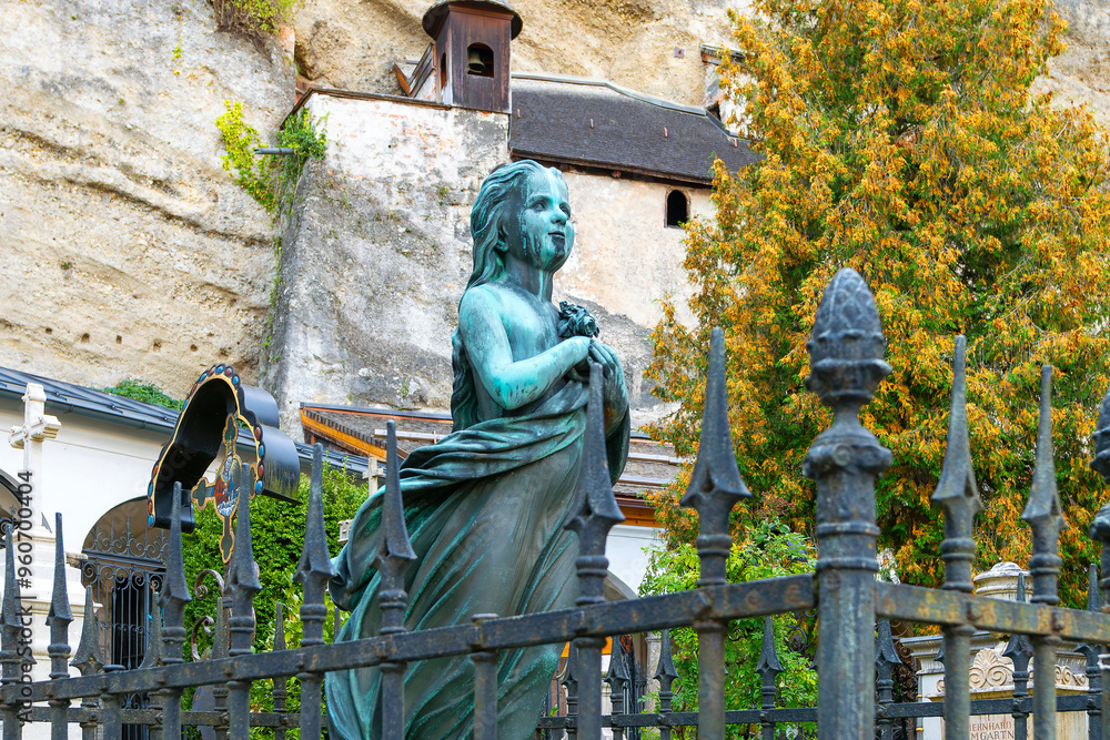 Obraz premium Bronze statue of a young girl as a memorial headstone at a grave in the St Peter's Cemetery, with the ancient abbey Catacombs behind, in Salzburg, Austria.