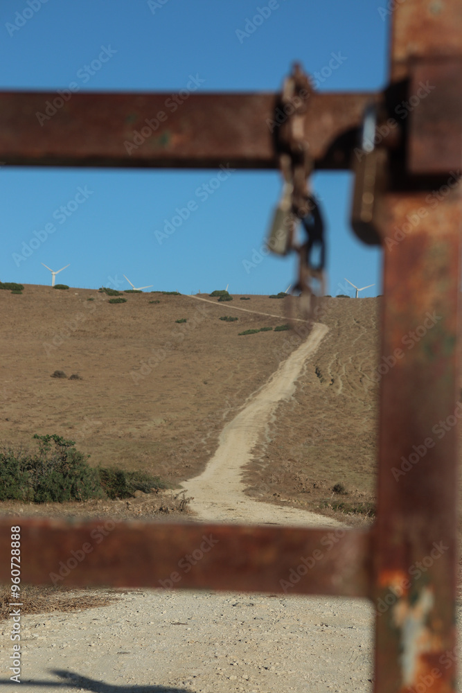 A sunny day with clear skies, there is a rusty metal gate on a field with a rusty lock on it. Being the gate one sees a gravel road going up the hill. A sunny day with clear skies, there is a rusty metal gate on a field with a rusty lock on it. Being the gate one sees a gravel road going up the hill.