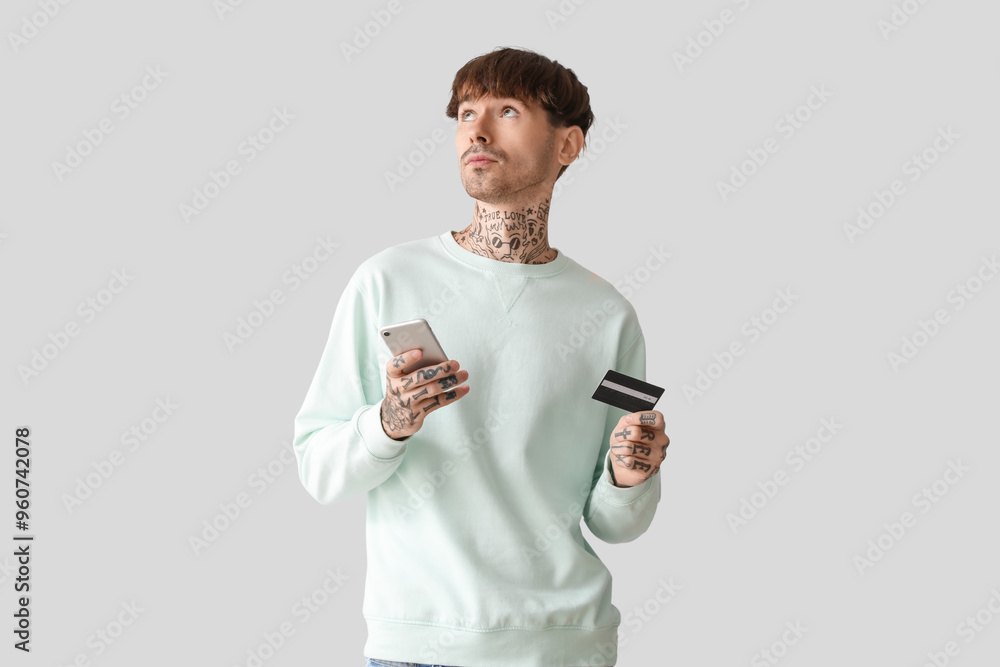 Tattooed young man with credit card and mobile phone on light background Tattooed young man with credit card and mobile phone on light background
