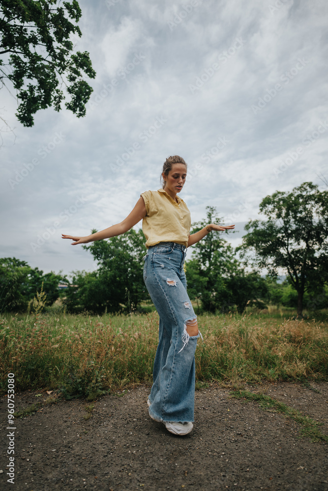 Fototapeta premium Young woman posing confidently outdoors in a casual outfit with ripped jeans and a yellow top, set against a cloudy sky.