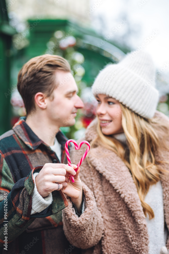 Obraz premium Beautiful young loving couple, boyfriend and girlfriend having fun on a christmas market wearing warm clothes. Outdoors, winter time, snowy weather. Tangerines in string bag, fir tree branches