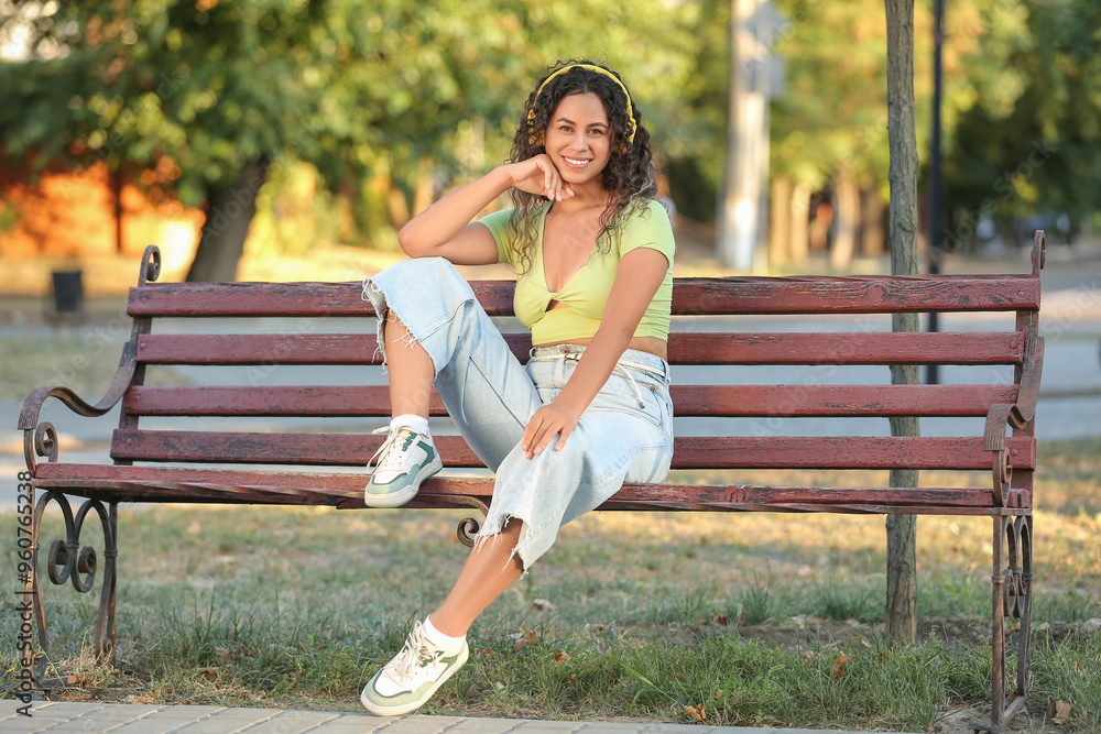 Fototapeta premium Happy young African-American woman in headphones sitting on bench in park