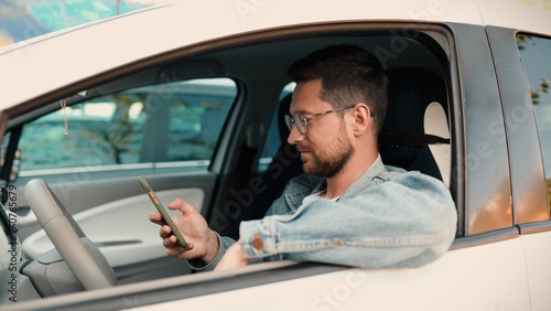 Close up of handsome young male texting on smartphone then looking at camera and smiling while sitting in white car. Happy Caucasian man tapping on cell phone. Driver seating in auto. Ride concept photo