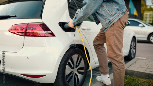 Close up of young man putting cable in charger port to charge his electric car outdoors. Male hand plugs in charger cable in vehicle at charger station and walks away. EV future technology. Eco car photo