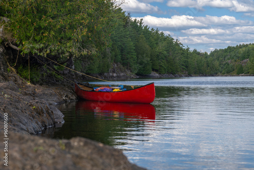 Bright red canoe loaded with camping gear tied to a tree by rugged rocky shore at French River Provincial park, Ontario, Canada. Camping, portaging, adventure, summer activities concept.