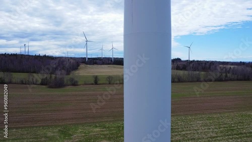 Un drone remonte lentement un mat d'éolienne jusqu'a son rotor situé au sommet avec en arrière-plan des champs cultivés, de la forêt et d'autres éoliennes en rotation sous un ciel bleu nuageux. photo