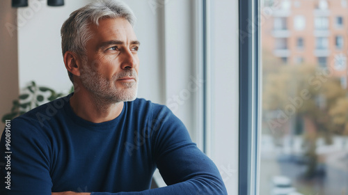 Attractive mature man leaning on the sill of a large bright window with copy space looking away