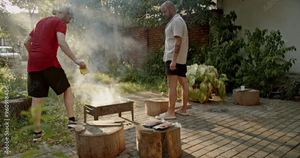 Two male friends in a yard are preparing a barbecue. They are gathering around the grill, putting oil on the meat, and getting ready to grill. Two male friends in a yard are preparing a barbecue. They are gathering around the grill, putting oil on the meat, and getting ready to grill.