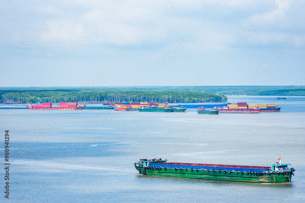 Obraz premium Barges and small boats navigate the flowing waters of the Mekong River in Vietnam, with a backdrop of lush tropical forest. 