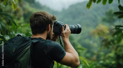 A man holds a camera, focused on photographing wildlife amidst the dense greenery of a rainforest on a foggy morning. The surrounding foliage adds to the serene atmosphere