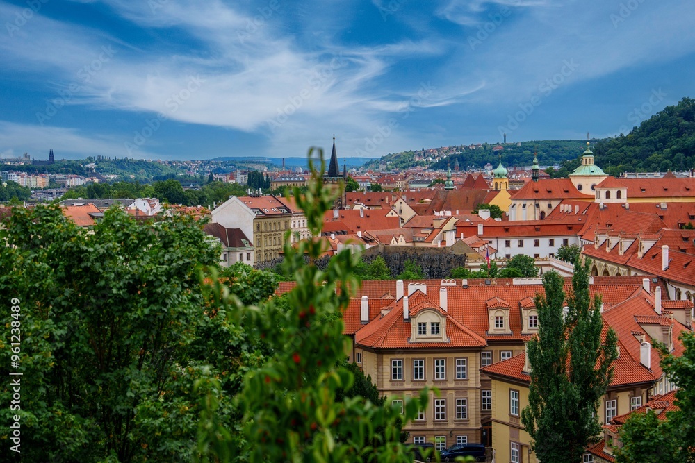 Obraz premium This beautiful cityscape of Prague features the iconic red-tiled roofs of the historic old town, framed by lush green trees under a bright blue sky. A perfect image for travel and tourism.