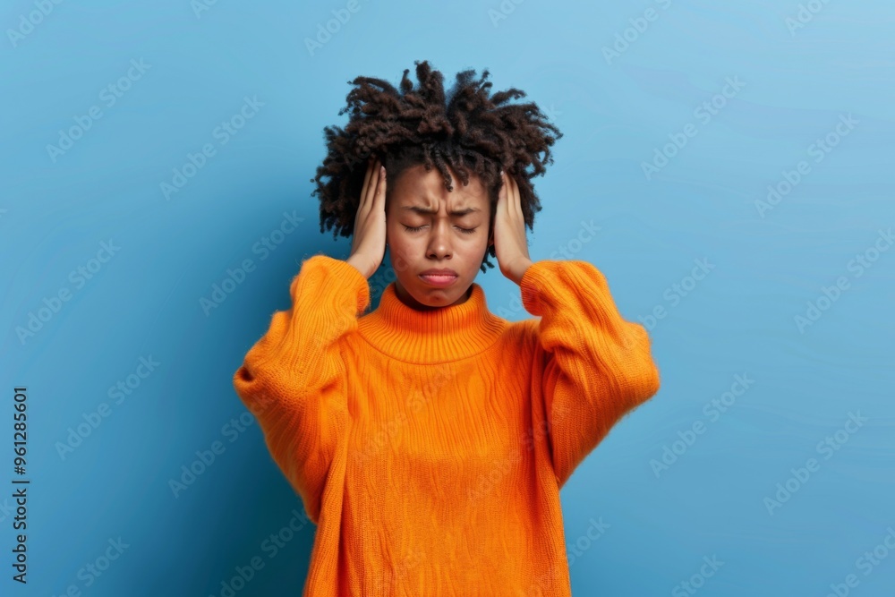 Frustrated young African American woman struggles with a migraine indoors after a noisy party while dressed in an orange sweater Frustrated young African American woman struggles with a migraine indoors after a noisy party while dressed in an orange sweater
