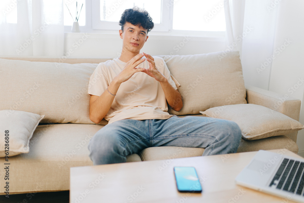 Young man sitting on a sofa in a bright room, casually dressed, with a phone and laptop on the table Relaxed atmosphere, modern interior design, natural light coming through windows Young man sitting on a sofa in a bright room, casually dressed, with a phone and laptop on the table Relaxed atmosphere, modern interior design, natural light coming through windows