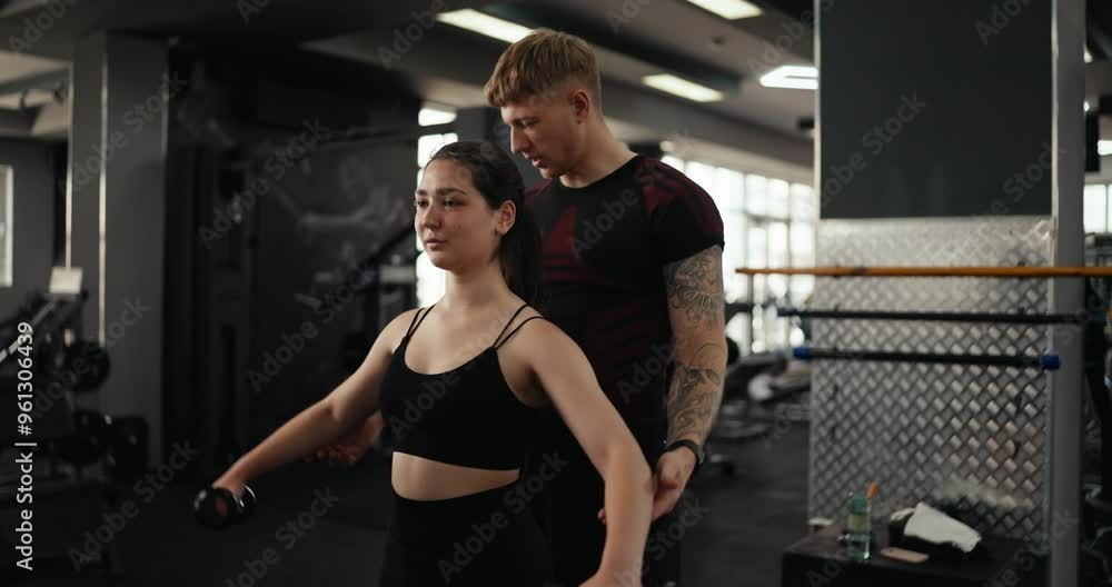 A confident guy a blond trainer in a black sports uniform and with tattoos supports a brunette girl and tells her how to correctly perform arm movements for effective exercise in the gym A confident guy a blond trainer in a black sports uniform and with tattoos supports a brunette girl and tells her how to correctly perform arm movements for effective exercise in the gym