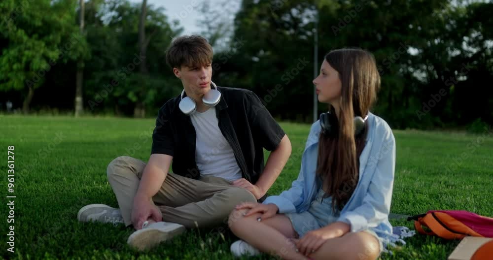 A teenage guy in wireless headphones communicates with his teenage girlfriend during their vacation and picnic on the green lawn in the park
