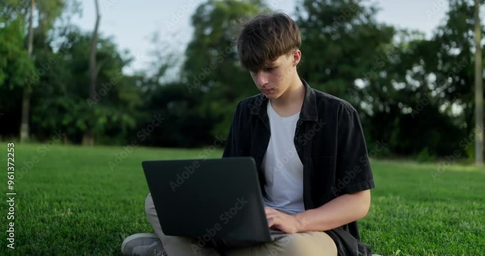 Focused teenage guy is engaged in self-development and typing on a laptop while sitting on the lawn in the park Focused teenage guy is engaged in self-development and typing on a laptop while sitting on the lawn in the park