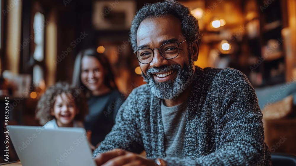 A smiling person works on their laptop while family members share a joyful moment behind them A smiling person works on their laptop while family members share a joyful moment behind them