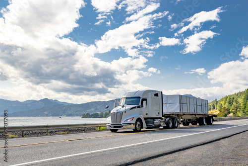 Bonnet powerful white big rig semi truck transporting covered fastened cargo on flat bed semi trailer running on the road along the railroad and river