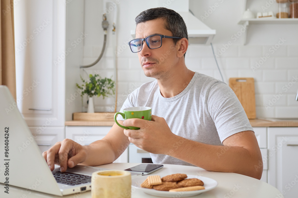 Serious focused man freelancer working from home in kitchen with laptop browsing internet using technology concentrated on work and career tasks enjoying coffee during remote work Serious focused man freelancer working from home in kitchen with laptop browsing internet using technology concentrated on work and career tasks enjoying coffee during remote work