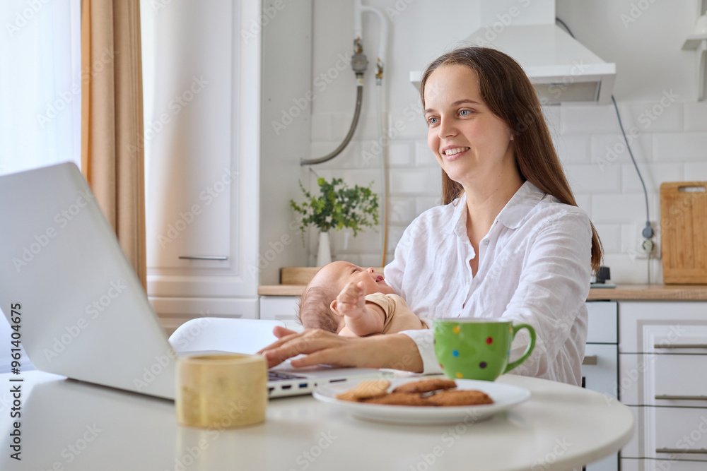 Smiling woman parent working from home on laptop busy with e-commerce job while cuddling newborn and managing online service sitting at table in home kitchen interior Smiling woman parent working from home on laptop busy with e-commerce job while cuddling newborn and managing online service sitting at table in home kitchen interior