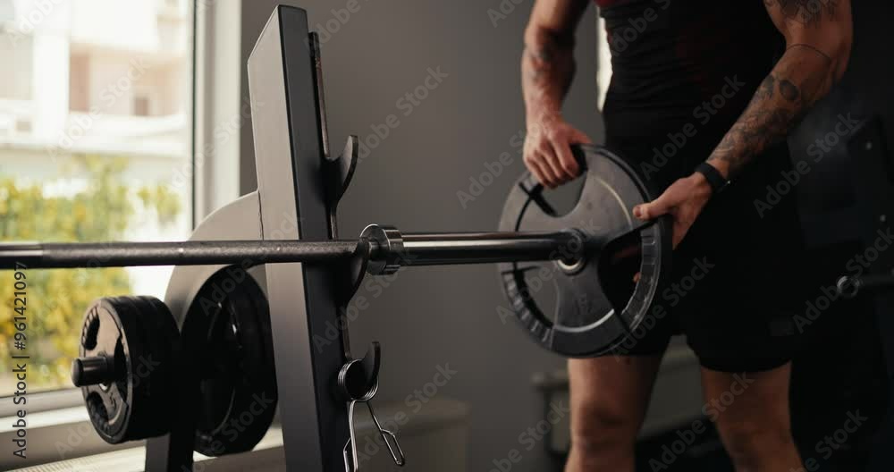 Athlete guy in a black sports uniform with tattoos puts weights on a barbell before starting his hard bench press and fitness training in the gym Athlete guy in a black sports uniform with tattoos puts weights on a barbell before starting his hard bench press and fitness training in the gym