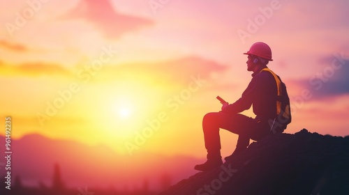 A silhouette of an engineer wearing a hard hat sits on a rock, looking out over a beautiful sunset. This image symbolizes hard work, determination, success, and the beauty of nature.