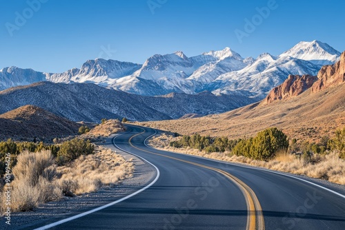 A sweeping wide-angle photo of a Nevada desert highway shows the road winding through desert scrub. Snow-capped mountains loom in the background, framed by a vibrant blue sky and clear horizon