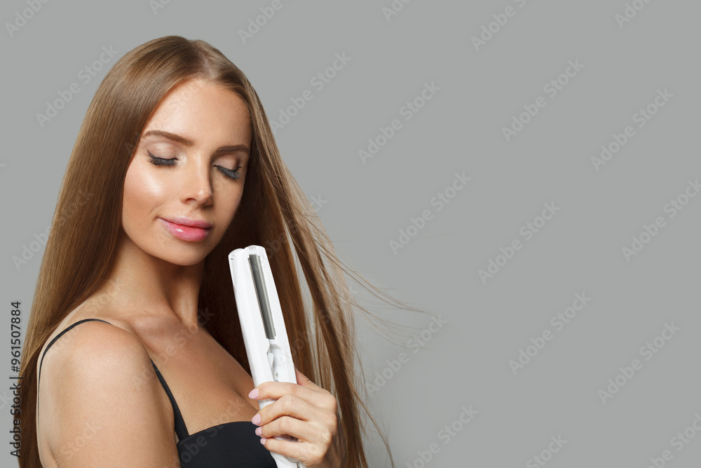 Joyful fashion model with brown shiny straight haistyle portrait. Cute woman with straightening iron in her hand on gray studio background Joyful fashion model with brown shiny straight haistyle portrait. Cute woman with straightening iron in her hand on gray studio background