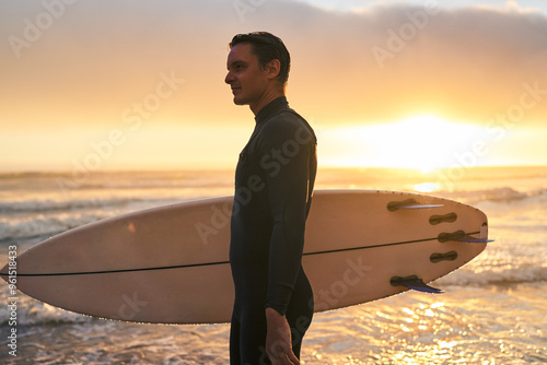 Slika na platnu A surfer in a wetsuit carrying a surfboard while standing on the beach during a beautiful sunset