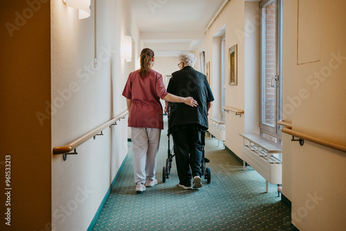 Rear view of female caregiver assisting senior woman walking with mobility walker in corridor